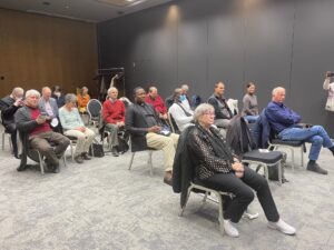 Teilnehmende sitzen in Stuhlreihen und blicken in Richtung der Redenden