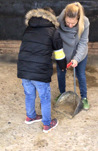 Ein Kind in Jacke und mit einer gelben Armbinde ist von hinten zu sehen. Eine Frau hilft beim Einsammeln der Pferdeäpfel 